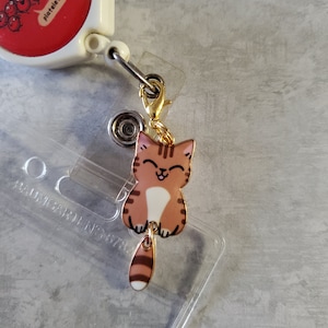 May include: A gold-coloured retractable badge holder with a red and white cartoon image of platelets. Attached to the holder is a charm of a brown and white cartoon cat with a gold chain.