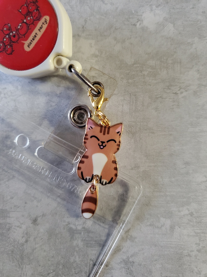 May include: A gold-coloured retractable badge holder with a red and white cartoon image of platelets. Attached to the holder is a charm of a brown and white cartoon cat with a gold chain.