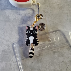 May include: A black and white cat charm with a pink nose and a gold chain. The charm is attached to a clear plastic badge holder with the text "UNICORN DESIGNS" printed on it.