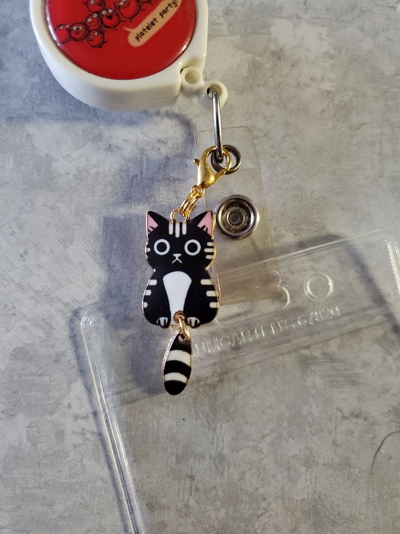 May include: A black and white cat charm with a pink nose and a gold chain. The charm is attached to a clear plastic badge holder with the text "UNICORN DESIGNS" printed on it.