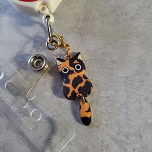 May include: A gold-coloured retractable badge holder with a red and white circular charm that says "platelet party!" attached to a black and orange cat charm with large eyes.