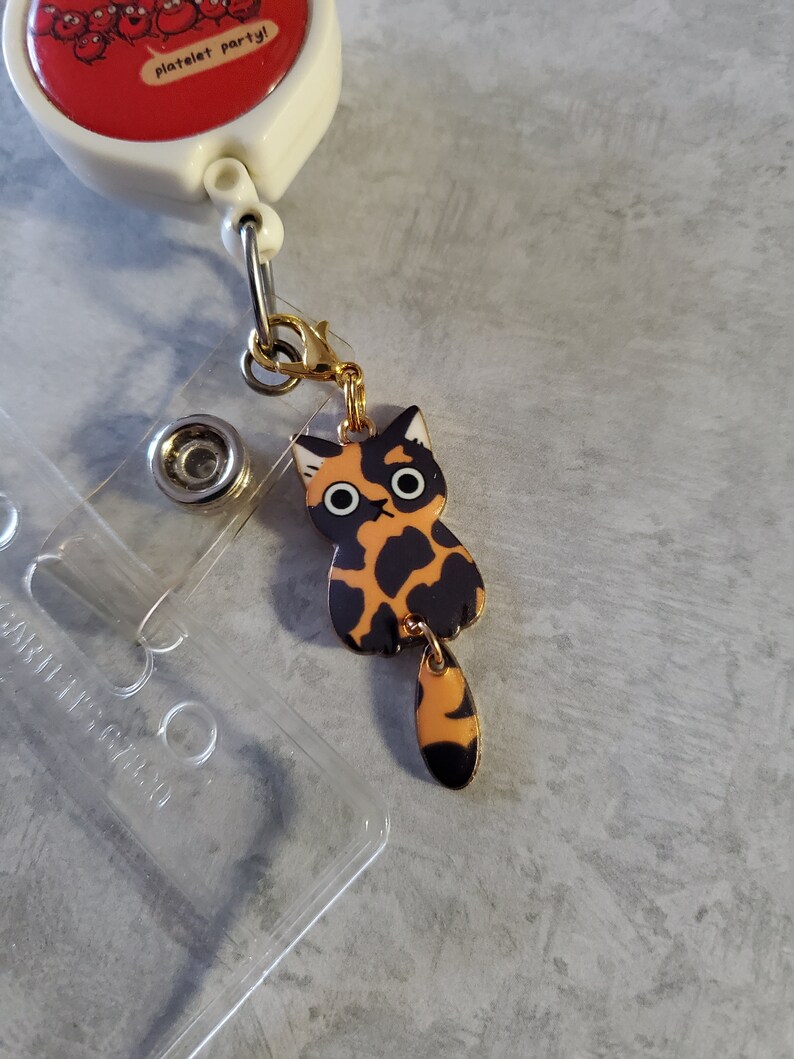 May include: A gold-coloured retractable badge holder with a red and white circular charm that says "platelet party!" attached to a black and orange cat charm with large eyes.