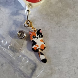 May include: A gold-coloured retractable badge holder with a red and white 'platelet party!' charm. A black, orange and white cat charm is attached to the holder.