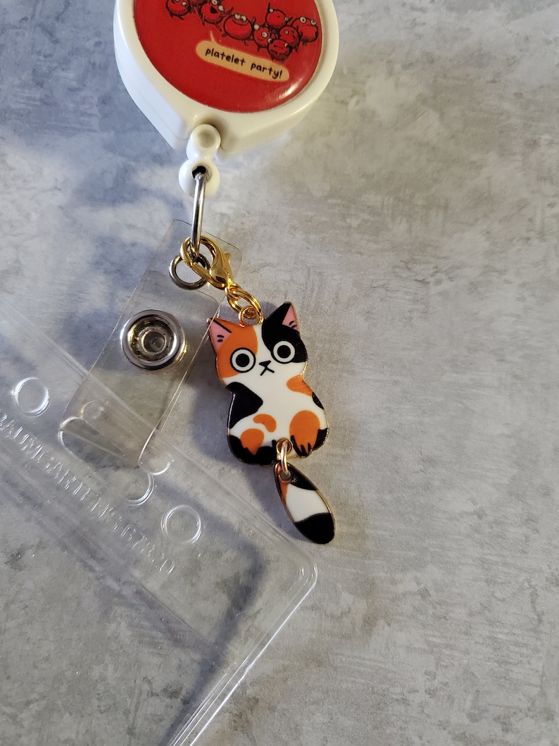 May include: A gold-coloured retractable badge holder with a red and white 'platelet party!' charm. A black, orange and white cat charm is attached to the holder.