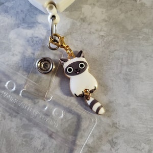 May include: A white and grey cat charm with a gold chain attached to a retractable badge holder. The badge holder has a clear plastic insert and the text "platelet party!" on the red circle.