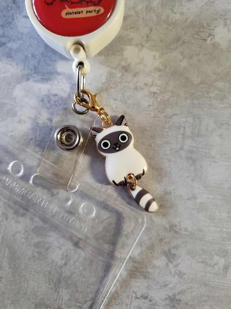 May include: A white and grey cat charm with a gold chain attached to a retractable badge holder. The badge holder has a clear plastic insert and the text "platelet party!" on the red circle.