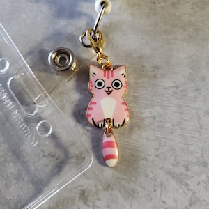 May include: A pink and white cat charm with a gold chain and a clip. The cat has pink stripes and a white belly. The charm is attached to a gold chain with a clip.
