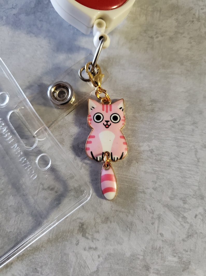 May include: A pink and white cat charm with a gold chain and a clip. The cat has pink stripes and a white belly. The charm is attached to a gold chain with a clip.