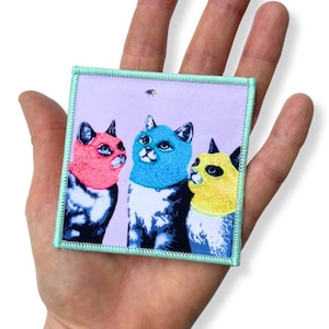 Könnte beinhalten: Quadratischer Aufnäher mit drei stilisierten Katzen mit farbenfrohem Fell und passenden Kragen. Die Katzen sind in Rosa, Blau und Gelb gehalten und vor einem lavendelfarbenen Hintergrund platziert. Der Aufnäher hat einen hellgrünen Rand.