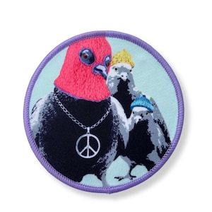 Könnte beinhalten: Gestickter Aufnäher mit drei stilisierten Tauben. Eine Taube hat einen roten Kopf und trägt eine silberne Friedenszeichen-Halskette. Die anderen beiden Tauben tragen gelbe und blaue Strickmützen. Der Aufnäher hat einen hellblauen Hintergrund und einen lila Rand.