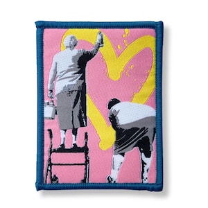 Könnte beinhalten: Gestickter Aufnäher mit einem Kunstwerk im Banksy-Stil, das zwei Figuren zeigt, die ein gelbes Herz auf rosa Hintergrund malen. Eine Figur steht auf einer Leiter, die andere beugt sich. Der Aufnäher hat einen blauen Rand.