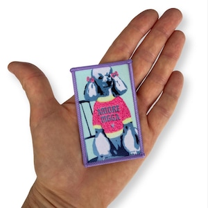Könnte beinhalten: Ein rechteckiger bestickter Aufnäher mit hellblauem Rand, der einen grauen Pudel in einem leuchtend pinken Pullover mit dem Text AMORE DIGGA in dunkelvioletten Buchstaben zeigt. Der Pudel trägt kleine rosa Schleifen an den Ohren und ein gelbes Halsband.