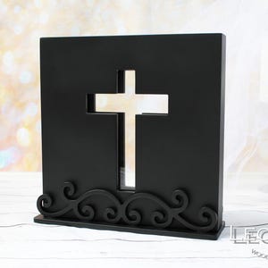 May include: A black wooden cross-shaped ornament with a cut-out cross design. The base has an ornate, swirling pattern. This decorative item is likely for religious or memorial purposes.