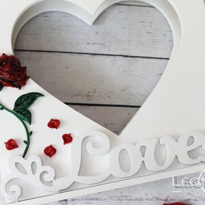 Könnte beinhalten: Weißer Holzrahmen in Herzform mit einer roten Rose und dem Wort "Love" in dekorativer Schrift. Der Rahmen hat eine ausgeschnittene Herzform und ist mit roten Rosenblättern verziert.