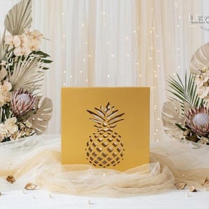 May include: A square, mustard-yellow box featuring a laser-cut pineapple silhouette on the front panel. The box is positioned centrally, displaying a clean, flat surface with a geometric cutout design that reveals the interior space through the shape.