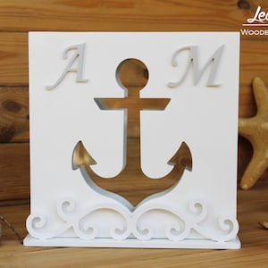 Könnte beinhalten: Weiße Holzkiste mit einem ausgeschnittenen Ankerdesign und den Buchstaben "A" und "M" auf beiden Seiten. Die Kiste hat ein dekoratives Schriftrollenmuster am Boden.
