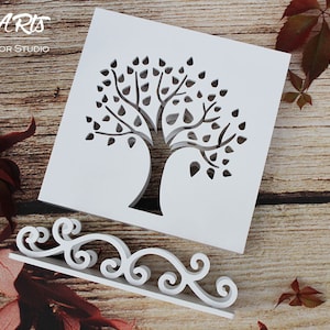 May include: White wooden cutout of a tree with leaves, on a white stand with a decorative scroll design. The stand and cutout are on a wooden surface with brown leaves.