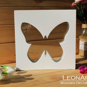 Könnte beinhalten: Weiße Holz-Wanddekoration mit einer ausgeschnittenen Schmetterlingsform. Der Schmetterling ist mittig auf dem weißen Quadrat platziert und hat einen Hintergrund aus hellem Holz.