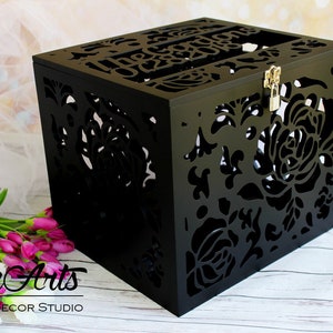 Puede incluir: Una caja de madera negra con intrincados patrones florales cortados con l&aacute;ser en los lados y la parte superior. La superficie superior presenta el texto "The Gardens 08.28.2016" cortado en la madera. Un peque&ntilde;o pestillo met&aacute;lico plateado cierra el frente.