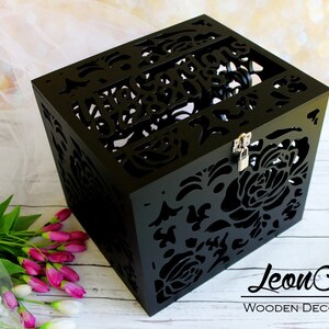 Puede incluir: Una caja cuadrada de madera negra con intrincados patrones florales cortados con l&aacute;ser en todos sus lados. La tapa incluye el texto recortado "The Gordons 07.28.2018" y un peque&ntilde;o pestillo de metal plateado con candado.