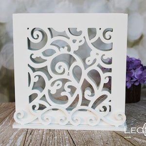 Könnte beinhalten: Weiße Holzkiste mit einem dekorativen Ausschnittmuster aus wirbelnden floralen Mustern. Die Kiste steht auf einer Holzoberfläche.