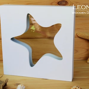 Könnte beinhalten: Weißer quadratischer Holzrahmen mit einem Ausschnitt in Form eines Seesterns. Der Rahmen befindet sich auf einem Holzgrund mit einem Seestern und Muscheln.