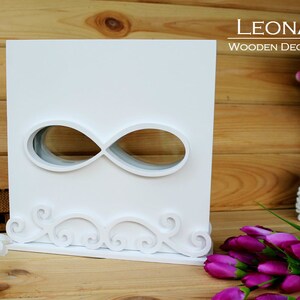Könnte beinhalten: Weiße Holzkiste mit einem Infinity-Symbol-Ausschnitt. Die Kiste hat unten ein dekoratives Schriftrollen-Design.  LEONARTS WOODEN DECOR STUDIO ist oben rechts auf der Kiste aufgedruckt.
