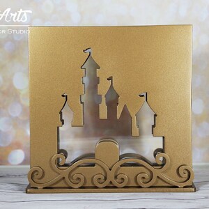 Könnte beinhalten: Eine goldene Holzbox in Form eines Schlosses mit einem dekorativen Schriftrollenmuster am Boden. Die Schlosssilhouette ist aus der Vorderseite ausgeschnitten und zeigt ein klares Fenster.