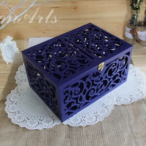 May include: A rectangular purple wooden box featuring intricate laser-cut scrollwork patterns on the lid and sides. The box includes a small gold-colored metal latch on the front, providing a secure closure for the hinged lid design.