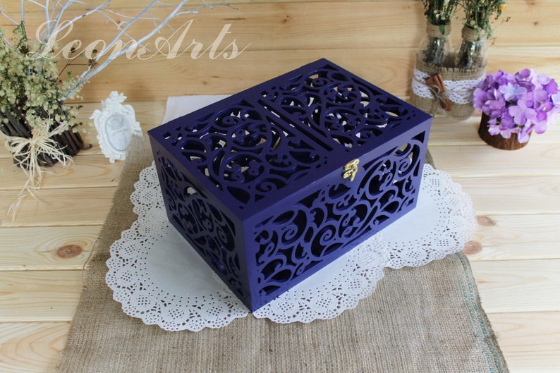 May include: A rectangular purple wooden box featuring intricate laser-cut scrollwork patterns on the lid and sides. The box includes a small gold-colored metal latch on the front, providing a secure closure for the hinged lid design.