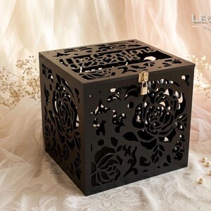 Puede incluir: Una caja cuadrada de madera negra con intrincados patrones florales cortados con l&aacute;ser en todos sus lados. La tapa superior presenta el texto The Gordons y 07.28.2018. Un peque&ntilde;o candado de metal plateado est&aacute; sujeto al cierre frontal.