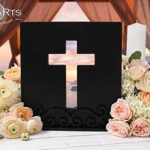 May include: A black wooden square plaque featuring a central cross-shaped cutout, positioned on a decorative base. The logo LEONARTS WOODEN DECOR STUDIO is visible in the top left corner. The cutout reveals a sunset view over the ocean.