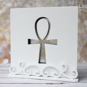 Könnte beinhalten: Weißes Holzdekor mit ausgeschnittenem Ankh-Symbol. Das Design umfasst dekorative Schnörkel am Sockel und den Text "LEONARTS Wooden Decor Studio" in der oberen rechten Ecke.