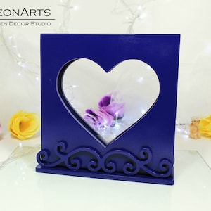 Könnte beinhalten: Ein dunkelblauer, herzförmiger Holzrahmen mit einem dekorativen Schriftrollenmuster am Boden. Der Rahmen ist leer und hat einen klaren Glaseinsatz.