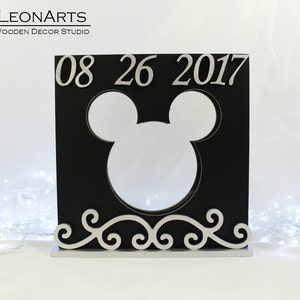 Könnte beinhalten: Schwarzer Holzrahmen mit einer Mickey-Maus-Silhouetten-Aussparung. Der Rahmen hat eine silberne Basis mit einem dekorativen Schriftrollen-Design und trägt das Datum "08 26 2017" in silbernen Lettern.