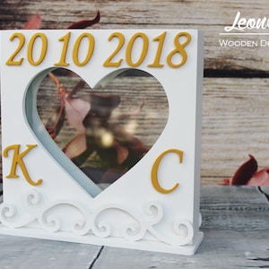 Könnte beinhalten: Weiße Holzkiste mit einem herzförmigen Ausschnitt und einer goldenen Inschrift mit der Aufschrift "2010 2018". Die Kiste hat auch die Buchstaben "K" und "C" in Gold auf beiden Seiten des Herzens.
