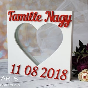 Könnte beinhalten: Weißer Holzrahmen mit einem herzförmigen Ausschnitt. Der Rahmen trägt den Text "Famille Nagy" und "11 08 2018" in roten Buchstaben. Der Rahmen steht auf einer Holzoberfläche.