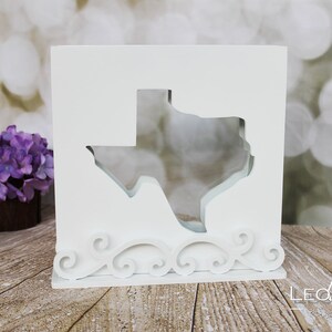 Könnte beinhalten: Weiße Holzkiste mit einem Ausschnitt des Bundesstaates Texas. Die Kiste hat unten ein dekoratives Schriftrollenmuster.