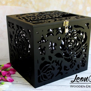 Puede incluir: Una caja cuadrada de madera negra con intrincados patrones florales cortados con l&aacute;ser en los lados y la parte superior. La caja incluye una ranura en la tapa, un pestillo de metal plateado y un peque&ntilde;o candado. El texto "Leon Arts WOODEN DECOR STUDIO" es visible.