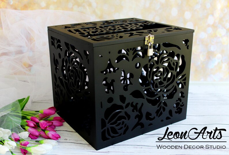 Puede incluir: Una caja cuadrada de madera negra con intrincados patrones florales cortados con l&aacute;ser en los lados y la parte superior. La caja incluye una ranura en la tapa, un pestillo de metal plateado y un peque&ntilde;o candado. El texto "Leon Arts WOODEN DECOR STUDIO" es visible.