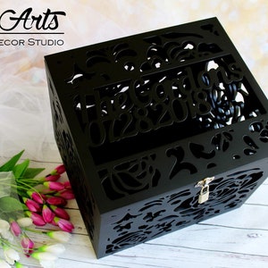 Puede incluir: Una caja de madera negra con intrincados patrones florales cortados con l&aacute;ser en todos sus lados. La parte superior presenta el texto "The Gardons" y "07282018" recortado en la madera. Un peque&ntilde;o candado de metal plateado est&aacute; en el cierre frontal.