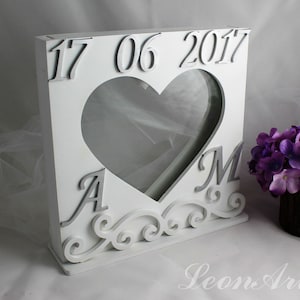 Könnte beinhalten: Weiße Holzkiste mit herzförmiger Öffnung und Glasscheibe. Die Kiste trägt das Datum "17 06 2017" und die Initialen "A & M" sind eingegraben. Die Kiste ist mit einem Rollendesign verziert.