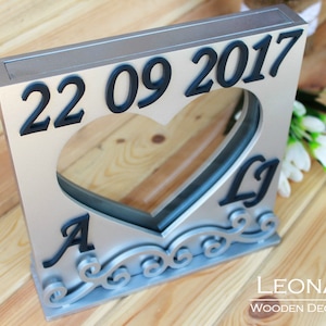 Könnte beinhalten: Eine silberne Holzkiste mit einer herzförmigen Öffnung und dem eingravierten Datum "22 09 2017" auf der Vorderseite. Die Kiste hat ein dekoratives Design um die Herzöffnung herum und die Initialen "A & L" sind im Herzen sichtbar.