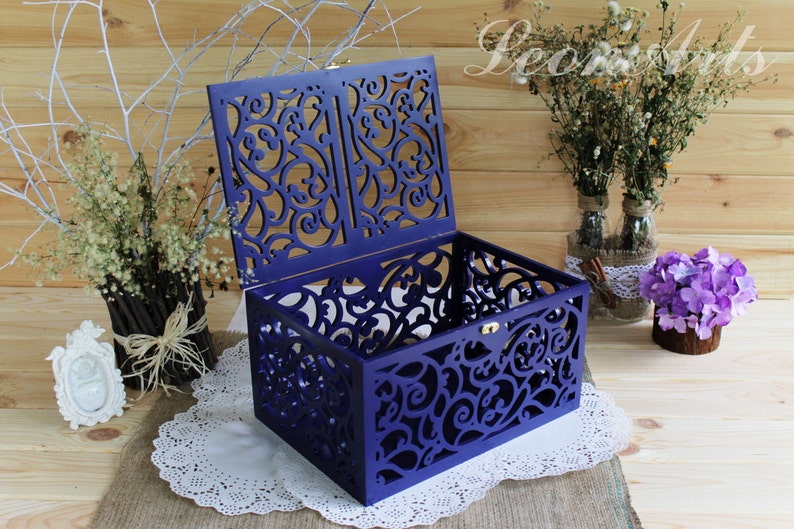May include: A rectangular wooden box in a deep purple hue featuring intricate laser-cut scrollwork patterns on all four sides and the hinged lid. The box includes a small gold-toned metal latch on the front edge to secure the lid when closed.