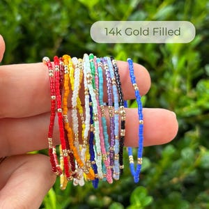 May include: A collection of colorful beaded bracelets with gold-filled accents. The bracelets feature a variety of colors, including red, yellow, green, blue, and black. The text "14k Gold Filled" is displayed in a white oval.