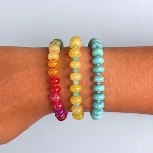 May include: Three beaded bracelets featuring smooth, round, disc-shaped beads. The left bracelet displays a gradient of green, yellow, orange, red, and purple. The center bracelet is yellow with small green spacers. The right bracelet is turquoise blue.