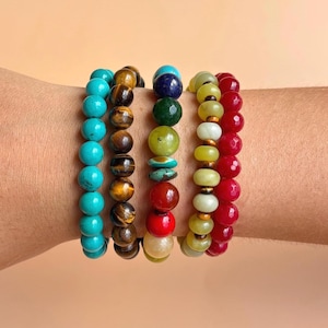 May include: Four beaded bracelets worn together. One features round turquoise beads, another has brown tiger eye beads, a third displays a mix of multicolored stones including blue, green, yellow, and red, and the fourth has yellow and red beads.