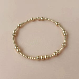 May include: A gold beaded bracelet with a mix of small and larger round beads. The bracelet is on a light pink background. The beads have a shiny finish and are strung together to form a flexible band.