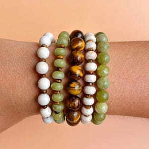 May include: A set of five beaded bracelets featuring various stones. One strand has matte white round beads, two strands have light green round beads, one strand has brown and gold tiger eye beads, and one strand has textured white beads.