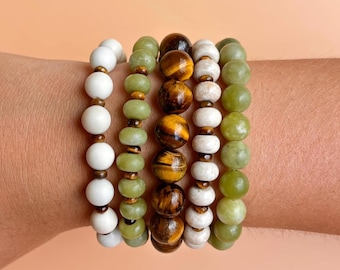 Puede incluir: Un conjunto de cinco pulseras de cuentas con varias piedras. Una hebra tiene cuentas redondas de color blanco mate, dos hebras tienen cuentas redondas de color verde claro, una hebra tiene cuentas de ojo de tigre marrón y dorado, y una hebra tiene cuentas blancas texturizadas.